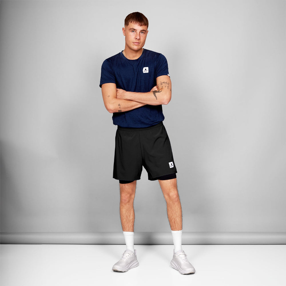 Man wearing a navy blue t-shirt and black shorts with a logo, standing against a gray background