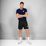 Man wearing a navy blue t-shirt and black shorts with a logo, standing against a gray background