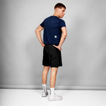 Man wearing a navy t-shirt and black shorts with a logo on a gray background